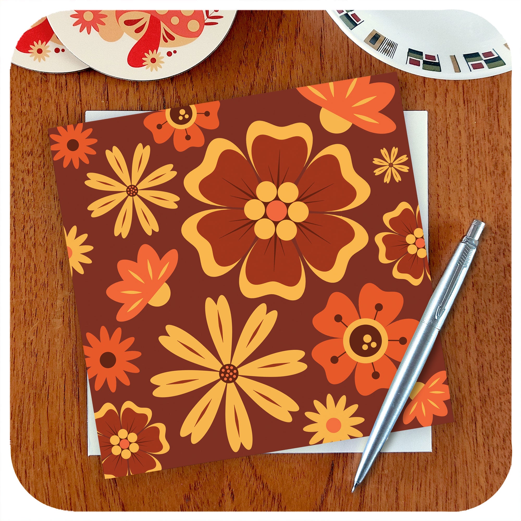 A greetings card with 70s style brown, orange and yellow flowers lies on a teak table with a silver pen | The Inkabilly Emporium