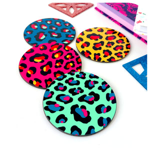 Four colourful Leopard Print Coasters lie scattered on a white table with books and glittery acrylic shapes in the background | The Inkabilly Emporium