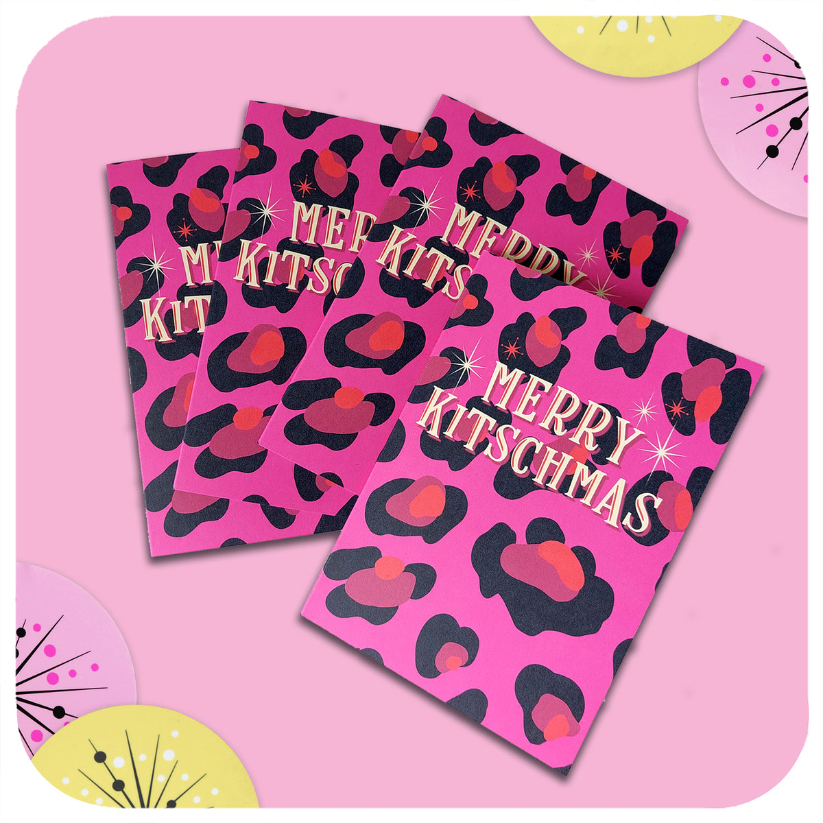 FOUR PINK LEOPARD PRINT CHRISTMAS CARDS LIE ON A PINK TABLE WITH 50S STYLE CHRISTMAS DECORATIONS. TEXT ON THE CARDS READS: MERRY KITSCHMAS.