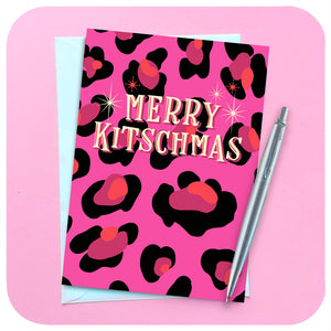 A pink leopard print christmas card lies on a pink background with a silver pen and white envelope. Text on the card reads: Merry Kitschmas.