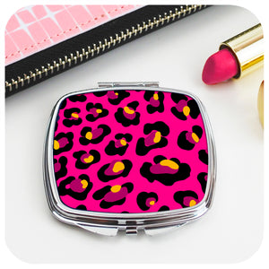 A square, chrome compact mirror with a pink leopard print design sits on a white table with an open pink lipstick and a pink purse.