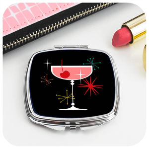 A Chrome compact mirror with a 50s style cocktail image sits on a white table with a lipstick and a pink purse in the background / The Inkabilly Emporium